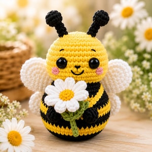 Könnte beinhalten: Ein gehäkeltes Bienen-Spielzeug mit einem runden, gelb-schwarz gestreiften Körper, großen schwarzen Augen, zwei schwarzen Fühlern und weißen Flügeln. Die Biene hält eine kleine weiße Margerite mit gelber Mitte und grünem Stiel in den Armen.