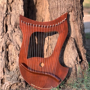 May include: A wooden lyre, stained a deep reddish-brown, leaning against a tree. The instrument has a curved shape, a sound hole, and many strings. The lyre is a stringed musical instrument, often used in ancient times.