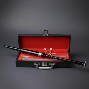 Puede incluir: Una gaita negra con detalles plateados, presentada en un estuche negro abierto con interior rojo. La funda contiene cañas y otros accesorios. El estuche tiene un asa y cierre plateados, sobre un fondo gris.