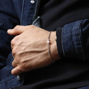 May include: A silver-colored bracelet featuring a small cross pendant. The bracelet is made of a delicate chain and is worn on the wrist. The image shows the bracelet against a dark background, highlighting its simple design.