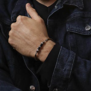Puede incluir: Una pulsera plateada con cuentas negras y plateadas. La pulsera se lleva en la muñeca, con una chaqueta vaquera oscura en el fondo. La pulsera tiene un diseño sencillo y elegante.