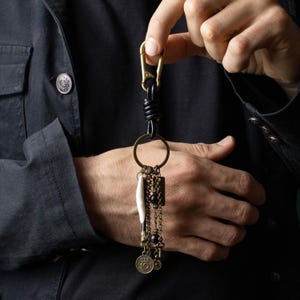 Könnte beinhalten: Ein handgefertigter Schlüsselanhänger mit einem Messingkarabiner und -ring. Der Schlüsselanhänger verfügt über eine schwarze Lederschlaufe, dekorative Ketten, Perlen und einen kleinen knochenartigen Anhänger. Zusätzliche Anhänger sind eine Münze und ein Schlüssel.