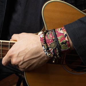 May include: A wrist adorned with multiple bracelets. One bracelet features a red, green, and black paisley pattern. Other bracelets include beads, a braided leather band, and a metal charm. The wrist rests on a wooden acoustic guitar.