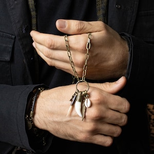May include: A bronze-toned necklace with a chain and a large ring holding various charms. Charms include a white tooth, a feather, a seashell, and a purple and white stone. The necklace is held in a person's hands.