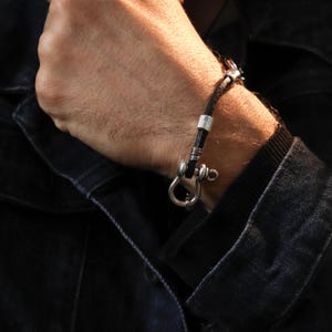 May include: A black leather bracelet with a silver-colored clasp and accents. The bracelet features a nautical-inspired design with a hook closure. The bracelet is worn on a person's wrist, against a dark denim jacket.