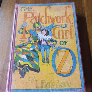 Może przedstawiać: Okładka starej książki dla dzieci zatytułowanej "Patchworkowa Dziewczynka z Krainy Oz" autorstwa L. Franka Bauma. Na okładce przedstawiono kolorową postać z patchworku trzymającą małą dziewczynkę w niebieskiej sukience i czapce.