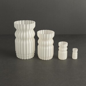 Könnte beinhalten: Vier weiße, gerippte Vasen unterschiedlicher Größe sind auf einer dunkelgrauen Oberfläche angeordnet. Die Vasen haben eine einzigartige, wellenförmige Form mit einem strukturierten, vertikalen Streifenmuster. Die kleinste Vase ist etwa 5 cm hoch, die größte etwa 20 cm.