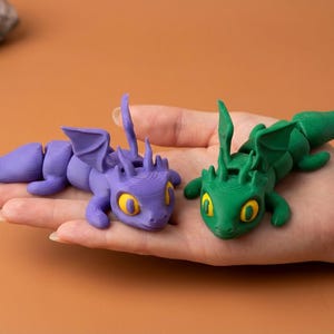 May include: Two articulated dragon figurines, one purple and one green, each with yellow eyes and detailed wings. The dragons are lying on a hand, showcasing their design and color contrast. The background is a solid orange.