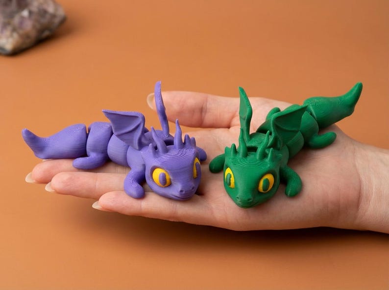 May include: Two articulated dragon figurines, one purple and one green, each with yellow eyes and detailed wings. The dragons are lying on a hand, showcasing their design and color contrast. The background is a solid orange.