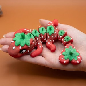 May include: A red and green strawberry-themed sculpture, resembling a dragon or lizard, is held in a hand. The sculpture has white dots and green leafy tops. The background is a solid orange color.