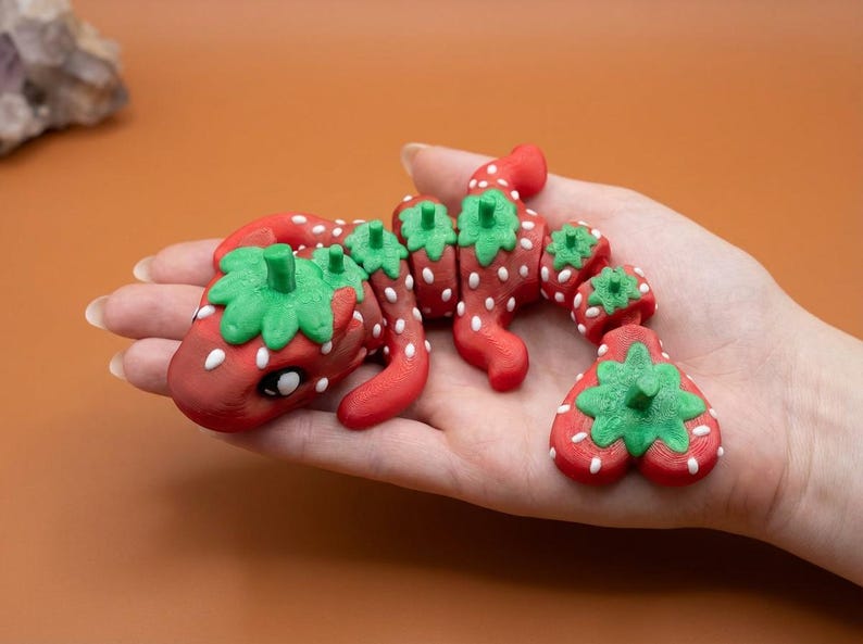May include: A red and green strawberry-themed sculpture, resembling a dragon or lizard, is held in a hand. The sculpture has white dots and green leafy tops. The background is a solid orange color.