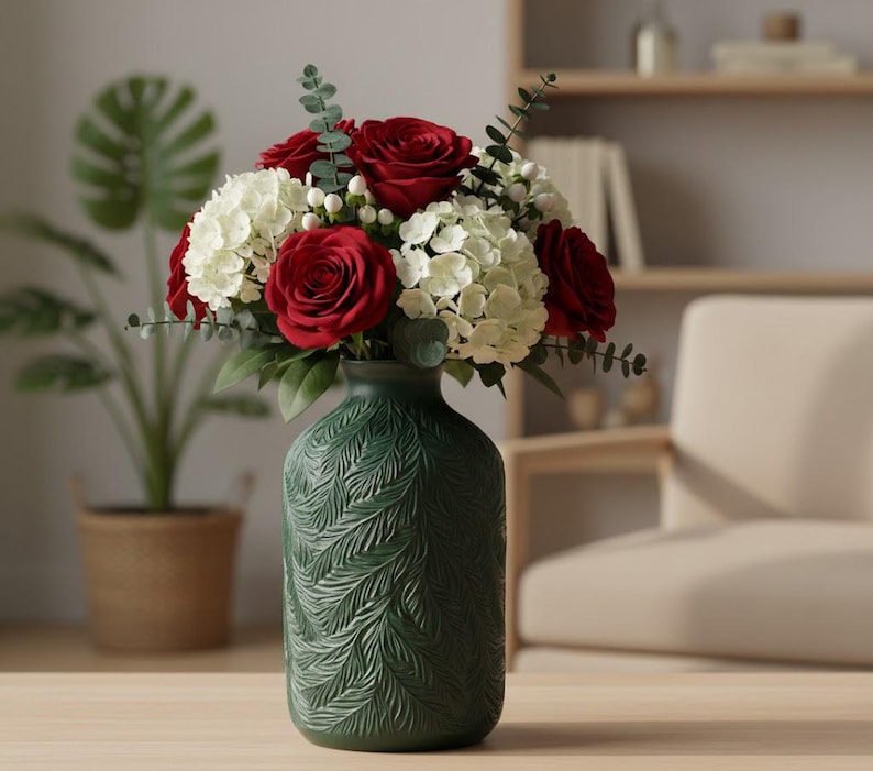 May include: A dark green ceramic vase with a textured, embossed fern leaf pattern on its exterior. The vase holds a floral arrangement consisting of deep red roses, white hydrangea clusters, small white berries, and green eucalyptus sprigs.