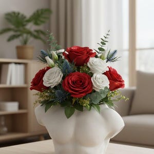 May include: A white ceramic vase shaped like a human torso filled with a floral arrangement of deep red roses, white roses, blue thistle, and green eucalyptus leaves. The vase has a smooth, matte finish and sits on a light wooden surface.