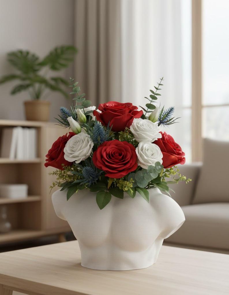 May include: A white ceramic vase shaped like a human torso filled with a floral arrangement of deep red roses, white roses, blue thistle, and green eucalyptus leaves. The vase has a smooth, matte finish and sits on a light wooden surface.