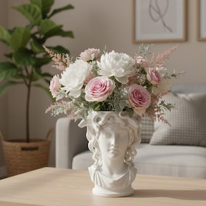 May include: A white ceramic vase shaped like a classical bust with snake-like hair, filled with an arrangement of white and pink flowers including roses, peonies, and feathery greenery. The vase features a detailed facial sculpture and draped neckline.
