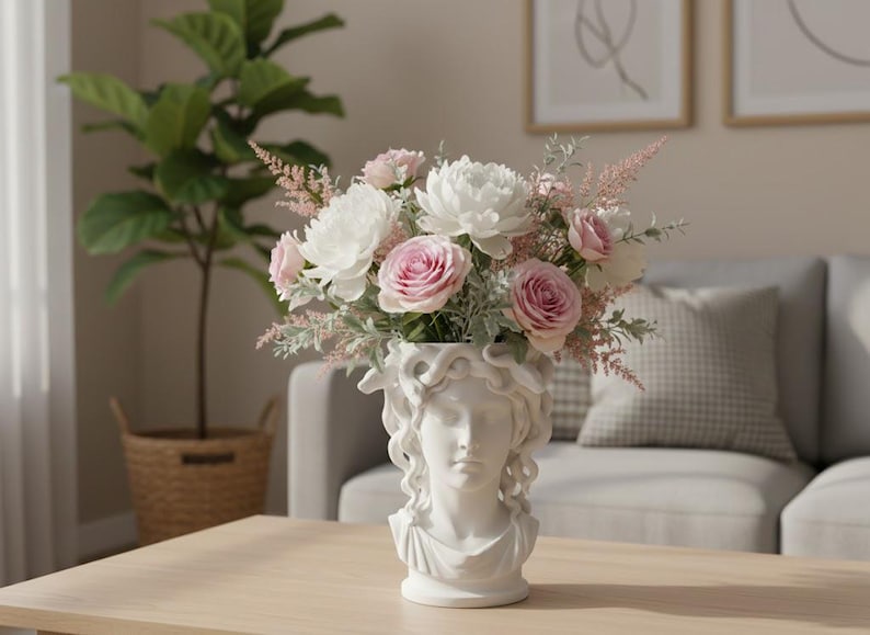 May include: A white ceramic vase shaped like a classical bust with snake-like hair, filled with an arrangement of white and pink flowers including roses, peonies, and feathery greenery. The vase features a detailed facial sculpture and draped neckline.