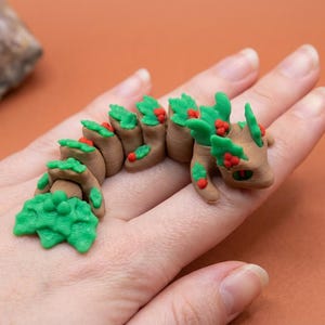 May include: A brown and green articulated dragon figurine with red berry accents. The dragon is made of clay and is posed on a hand. The dragon has green leafy sections and red berries.