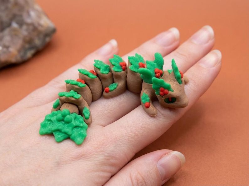 May include: A brown and green articulated dragon figurine with red berry accents. The dragon is made of clay and is posed on a hand. The dragon has green leafy sections and red berries.