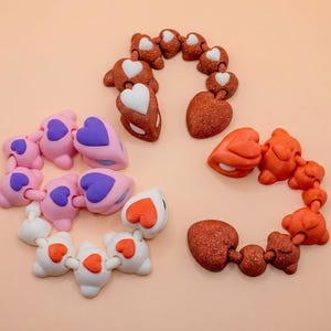 May include: An assortment of decorative heart and bear-shaped embellishments in various colors. The collection includes pink, white, and brown pieces, each with contrasting heart accents in purple, red, and white. These are likely for crafting or jewelry making.