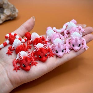 May include: A collection of small, colorful dragon figurines in red, white, and pink hues. The dragons have detailed features, including horns and scales. Some are curled, while others are posed. The figurines are held in a hand against an orange background.