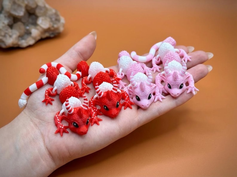 May include: A collection of small, colorful dragon figurines in red, white, and pink hues. The dragons have detailed features, including horns and scales. Some are curled, while others are posed. The figurines are held in a hand against an orange background.