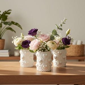 May include: Three white cylindrical vases featuring a raised heart pattern on the exterior. Each vase is filled with a floral arrangement containing light pink roses, white ranunculus, deep purple lisianthus, baby's breath, and green eucalyptus.