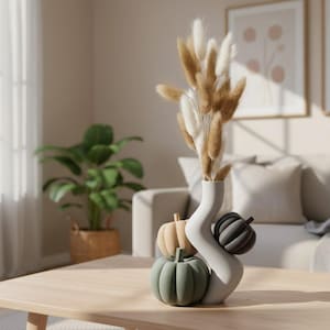 May include: A white, wavy-shaped ceramic vase holding a bouquet of dried, fluffy, tan and white bunny tail grass. The vase is decorated with three small, round, pumpkin-shaped ornaments in muted shades of sage green, beige, and dark gray.
