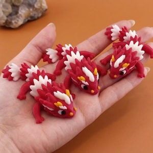 May include: Handful of miniature red and white dragon figurines with yellow accents. The dragons have detailed scales and are posed on a light brown surface. The figurines are small, detailed, and appear to be handmade.