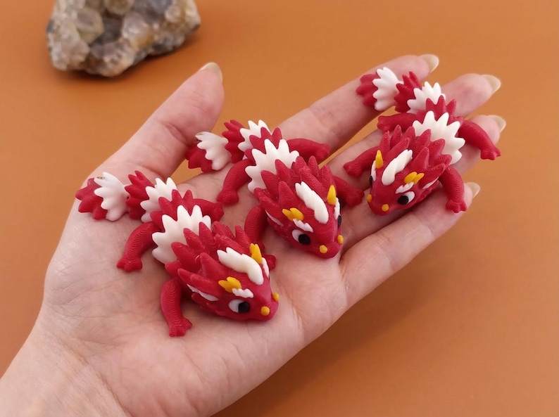May include: Handful of miniature red and white dragon figurines with yellow accents. The dragons have detailed scales and are posed on a light brown surface. The figurines are small, detailed, and appear to be handmade.