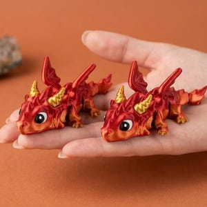 May include: Two miniature red and orange dragon figurines with gold horns and large eyes. The dragons are resting on a person's open palm, showcasing their detailed design. The background is a solid orange color.