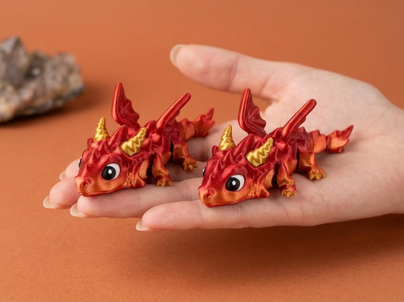 May include: Two miniature red and orange dragon figurines with gold horns and large eyes. The dragons are resting on a person's open palm, showcasing their detailed design. The background is a solid orange color.