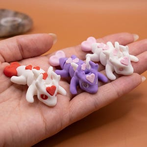 May include: A collection of small, handmade dragon figurines. The dragons are white and purple, with heart-shaped accents in red and pink. The figurines are lying in a person's hand.