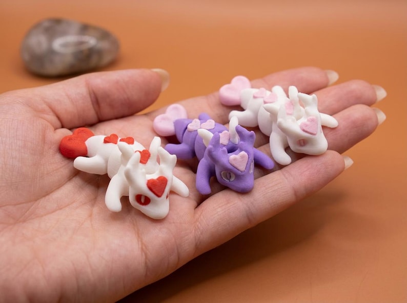 May include: A collection of small, handmade dragon figurines. The dragons are white and purple, with heart-shaped accents in red and pink. The figurines are lying in a person's hand.
