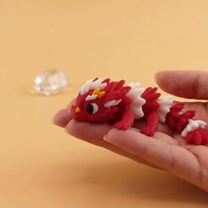 May include: A red, white, and yellow dragon figurine with a detailed design. The dragon is held in a hand against a light orange background. The dragon's body is red with white and yellow accents.
