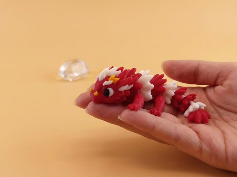 May include: A red, white, and yellow dragon figurine with a detailed design. The dragon is held in a hand against a light orange background. The dragon's body is red with white and yellow accents.