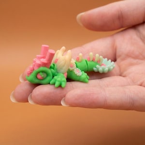 May include: A miniature, colorful sculpture of a fantastical creature, held in a hand. The sculpture features a green body, pink coral-like structures, and accents of white and light green. The overall design is whimsical and detailed.