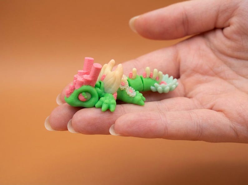 May include: A miniature, colorful sculpture of a fantastical creature, held in a hand. The sculpture features a green body, pink coral-like structures, and accents of white and light green. The overall design is whimsical and detailed.