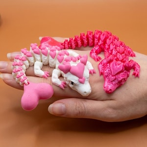 May include: Three 3D-printed dragon figurines. One is white with pink heart accents, another is solid pink, and the third is a pink and white dragon. The dragons are on a hand against a brown background.