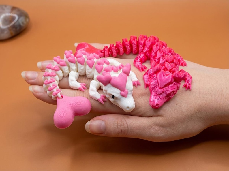 May include: Three 3D-printed dragon figurines. One is white with pink heart accents, another is solid pink, and the third is a pink and white dragon. The dragons are on a hand against a brown background.