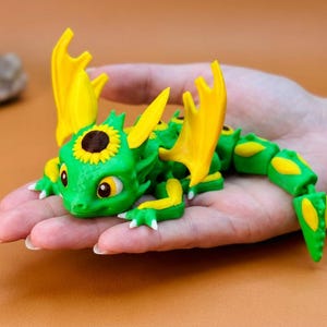 May include: A green and yellow articulated dragon figurine with sunflower accents. The dragon has large yellow wings and a sunflower on its head. The figurine is held in a hand, showcasing its details and design.