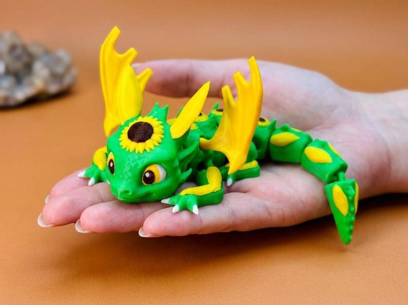 May include: A green and yellow articulated dragon figurine with sunflower accents. The dragon has large yellow wings and a sunflower on its head. The figurine is held in a hand, showcasing its details and design.