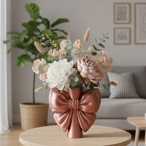 May include: A metallic rose gold vase shaped like a large bow, filled with a floral arrangement of white and dusty pink peonies, green eucalyptus leaves, and beige dried grasses, creating a soft and balanced decorative centerpiece.