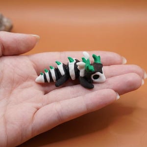 May include: A miniature, handmade dragon figurine. The dragon is black and white with green accents, and is approximately 2 inches long. The figurine is resting in a person's hand, with a blurred orange background.