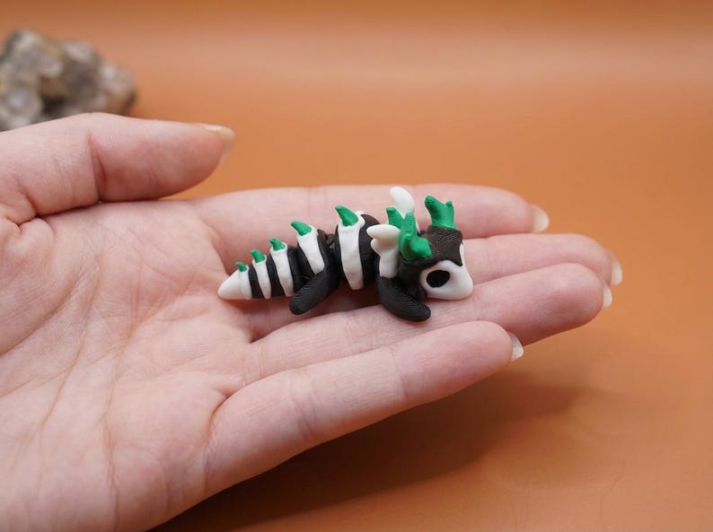 May include: A miniature, handmade dragon figurine. The dragon is black and white with green accents, and is approximately 2 inches long. The figurine is resting in a person's hand, with a blurred orange background.