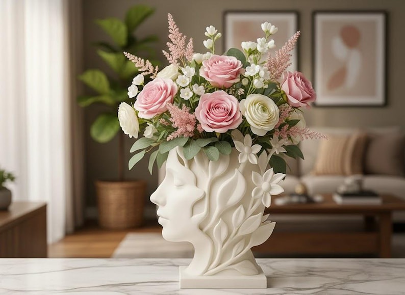 May include: A white ceramic vase shaped like a human head with closed eyes, adorned with relief carvings of leaves and flowers. The vase is filled with a vibrant bouquet of pink roses, white ranunculus, and feathery pink astilbe stems.
