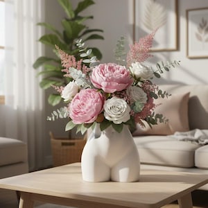 May include: A white ceramic vase shaped like a human torso holds a floral arrangement featuring large pink peonies, white roses, feathery pink astilbe, and sprigs of green eucalyptus leaves, creating a balanced and textured botanical display.