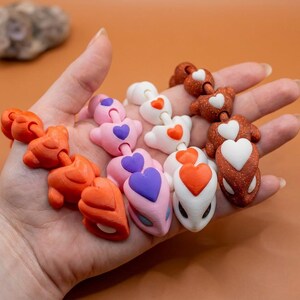 May include: A hand holds several colorful, connected heart-shaped charms. The charms are in shades of orange, pink, white, and brown, each with a contrasting heart detail. The background is a solid, warm orange.