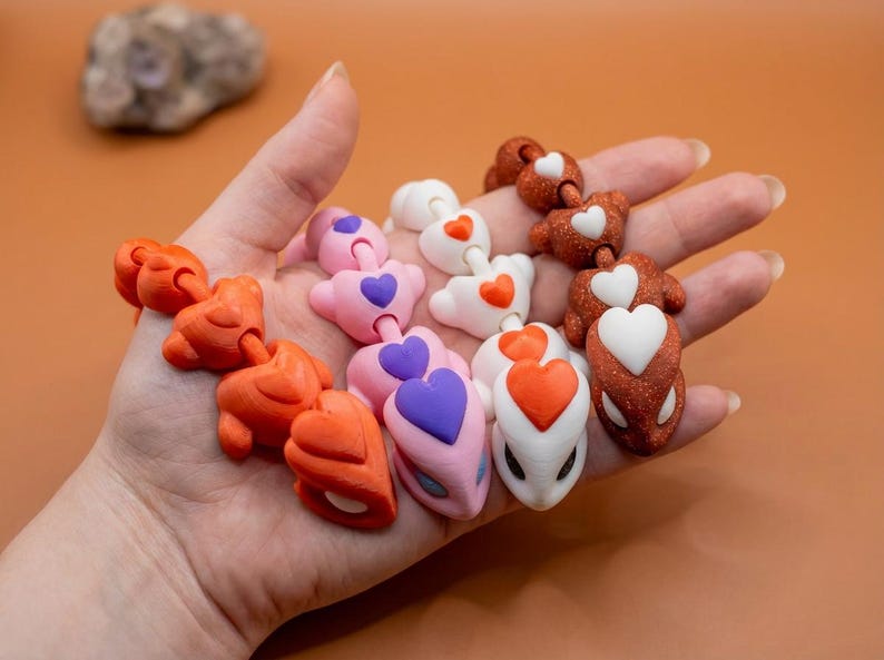 May include: A hand holds several colorful, connected heart-shaped charms. The charms are in shades of orange, pink, white, and brown, each with a contrasting heart detail. The background is a solid, warm orange.