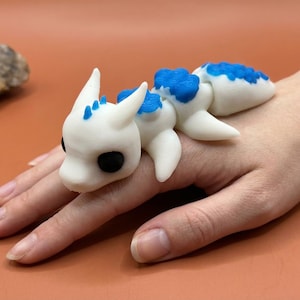 May include: A white and blue dragon figurine with black eyes. The dragon is lying on a hand. The figurine has a segmented body and is made of a smooth material. The dragon's back and head have blue accents.
