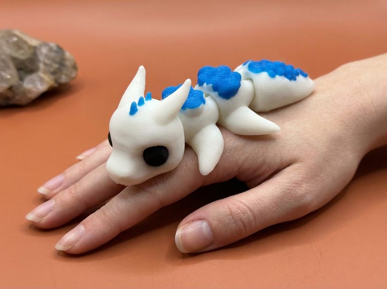 May include: A white and blue dragon figurine with black eyes. The dragon is lying on a hand. The figurine has a segmented body and is made of a smooth material. The dragon's back and head have blue accents.